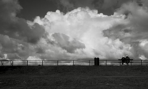 People on bench against cloudy sky