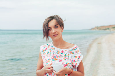 Portrait of woman holding coffee cup while standing against sea
