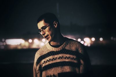 Young man wearing eyeglasses looking down while standing against sky at night