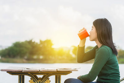 Young woman drinking coffee on table at sidewalk cafe by lake