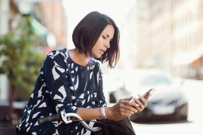 Businesswoman using smart phone while leaning on bicycle during summer