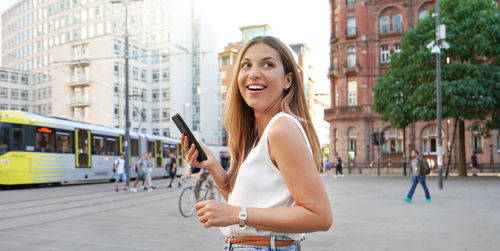 Attractive laughing woman holding smartphone walking on urban background. looking to the side. 