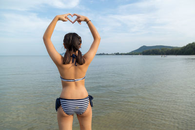 Rear view of woman making heart shape while standing in sea against sky