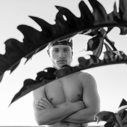 Low angle portrait of shirtless young man with arms crossed standing against sky