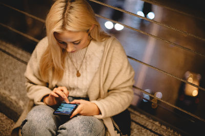 Young woman using smart phone while sitting on bridge in city at night