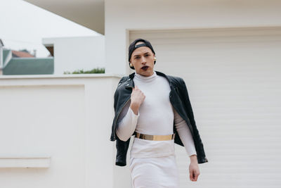 Portrait of transgender man wearing cap with jacket standing against building