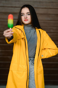 A girl in a yellow raincoat and a gray sweater holds a multicolored ice cream in her hand