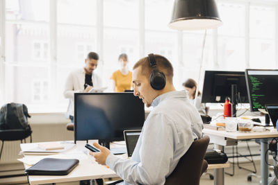 Side view of male engineer using mobile phone while coworkers working in background at creative workplace