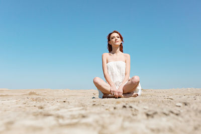 Young woman sitting on sand at beach against clear blue sky
