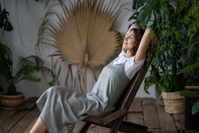 Young woman relaxed sit on comfortable armchair in stylish indoor garden with green exotic plants