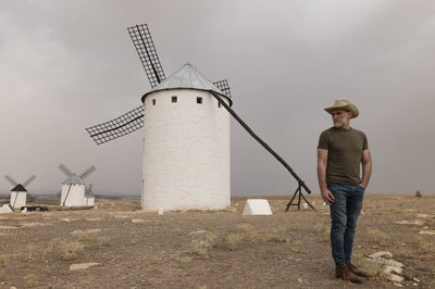 Man with cowboy hat standing by spanish traditional white windmills against sky