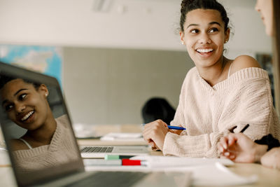 Smiling teenager studying with friend while sitting by table in classroom