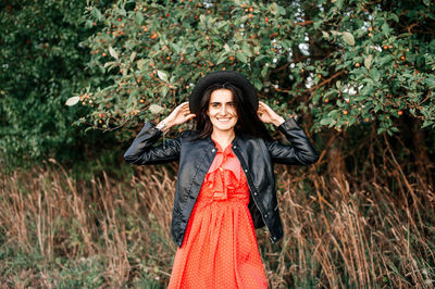 Portrait of a young woman standing against a background of plants in a red dress and a black hat