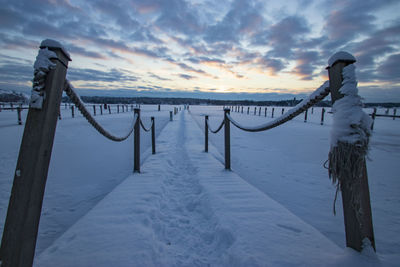 Wooden posts on snow covered railing against sky during sunset