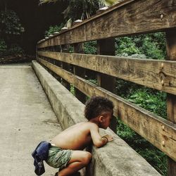 Side view of shirtless boy crouching by wooden railing