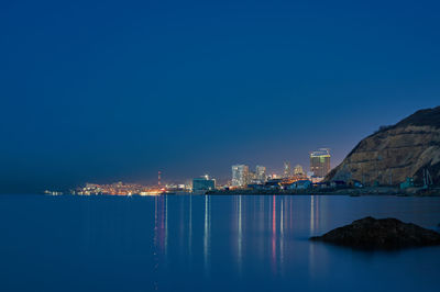 Illuminated city by sea against clear blue sky at night