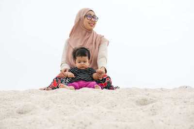 Mother with daughter sitting on sand at beach against clear sky