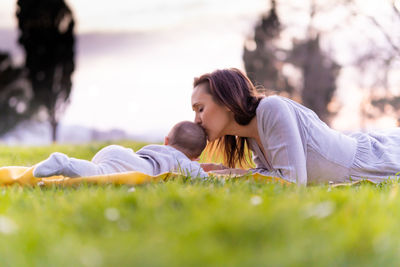 Side view of mother kissing baby while lying down on grass outdoors