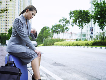 Side view of businesswoman checking time while sitting on luggage on sidewalk