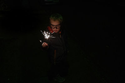 Cute boy holding illuminated sparkler while standing against black background