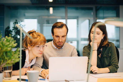 Female colleagues looking at businessman using laptop while sitting at desk in creative office