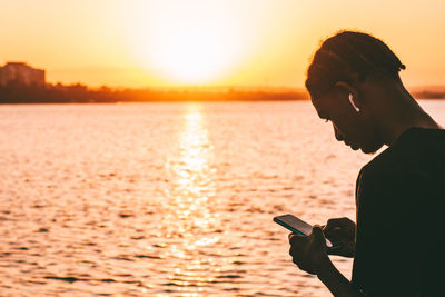 Side view of man photographing sea against sky during sunset