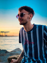 Portrait of young man wearing sunglasses while standing at beach against sky during sunset