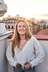 Cheerful attractive female in casual sweater standing on building rooftop and looking at camera with pretty smile in early evening