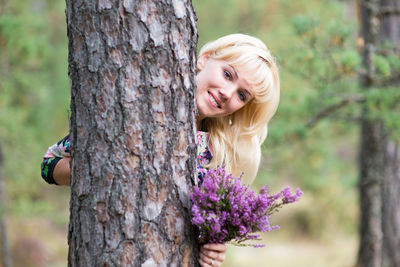 Portrait of smiling young woman holding flowers with tree trunk