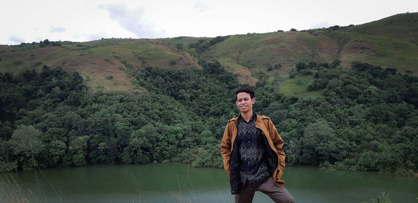 Portrait of young man standing by tree against sky