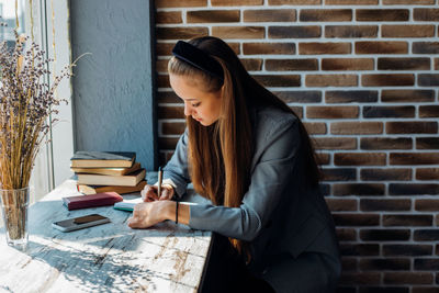 A young woman is sitting at a table by the window and writing in a notebook. 