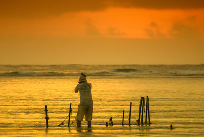 Rear view of fisherman in sea against orange sky during sunset