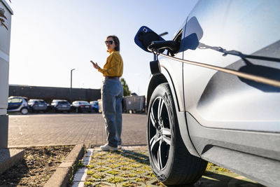 Electric car charging at station with woman in background holding smart phone on sunny day