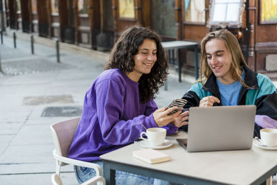 Two people using laptop and mobile phone while sitting outdoors at a coffee shop