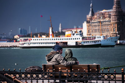 Rear view of couple sitting by river on bench in city