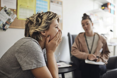 Depresses female student covering mouth with hand sitting near counselor in school office