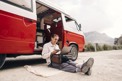 Smiling young man doing video call on laptop while sitting near van during vacation