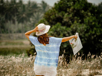 Woman wearing hat while holding map on field against trees