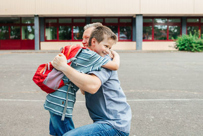 Happy young dad hugging his schoolboy son in front of the school building. 
