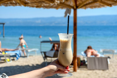 Personal perspective of person holding glass of cold iced coffee on beach in summer.