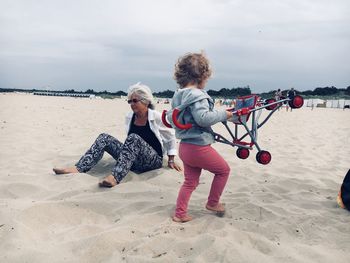 Full length of baby carrying stroller by woman at beach against sky