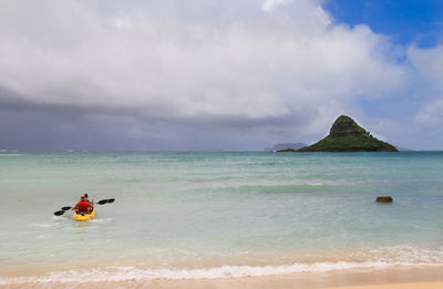 Rear view of people canoeing in sea against cloudy sky