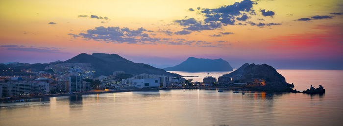 Sea by illuminated cityscape against sky at sunset