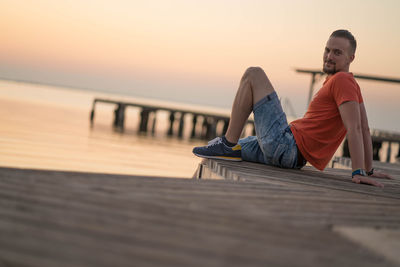 Man sitting on pier over sea against sky during sunset