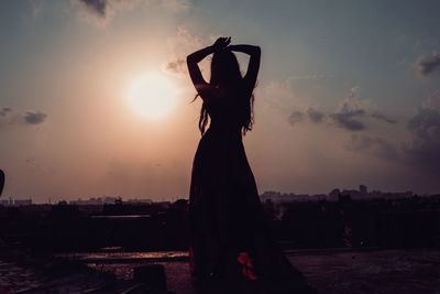 Silhouette woman with arms raised standing in city during sunset