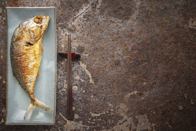 Large fried mackerel in rectangular ceramic plate beside chopsticks on rusty texture background