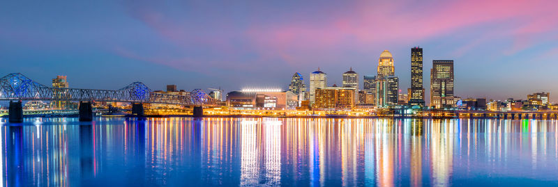 Illuminated buildings by river against sky at dusk