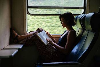 REAR VIEW OF YOUNG WOMAN SITTING BY WINDOW