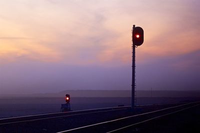 Signal by railroad tracks against sky at sunset