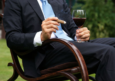 Midsection of businessman holding wine and cigar while sitting outdoors
